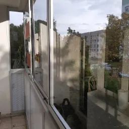 Zabudowany balkon z przesuwnymi oknami, widok na osiedle z drzewami, refleksy na szybach.