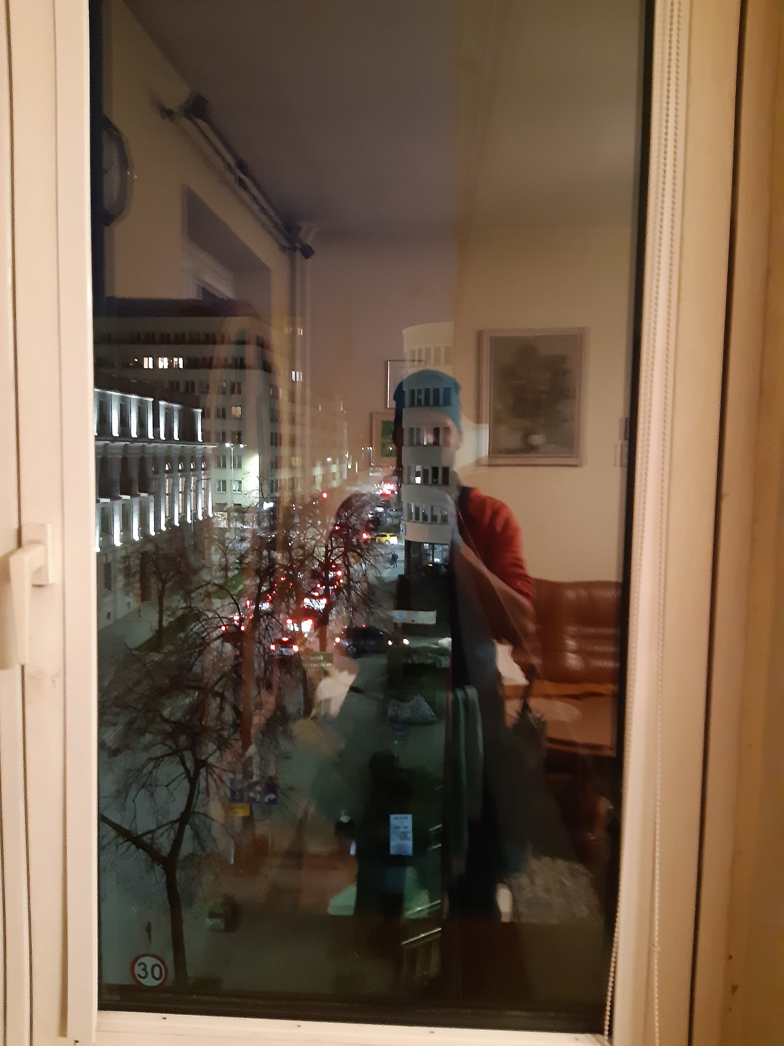 Odbicie osoby w nieumytym oknie, w tle widok na oświetlony budynek miejski i ulicę po zmroku.