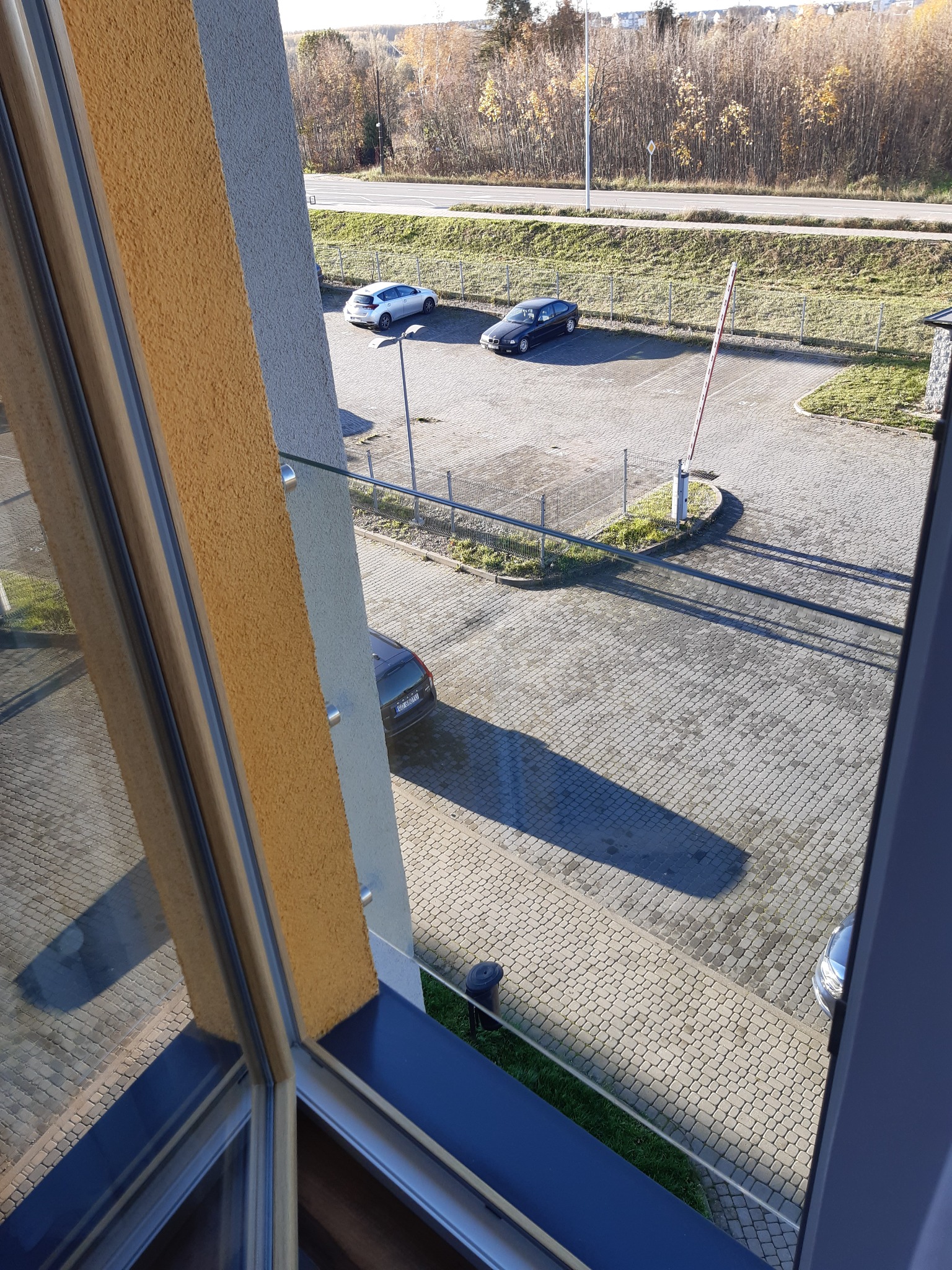 Widok z okna na parking z dwoma samochodami i szlabanem, z widocznym fragmentem ramy okiennej i parapetu.