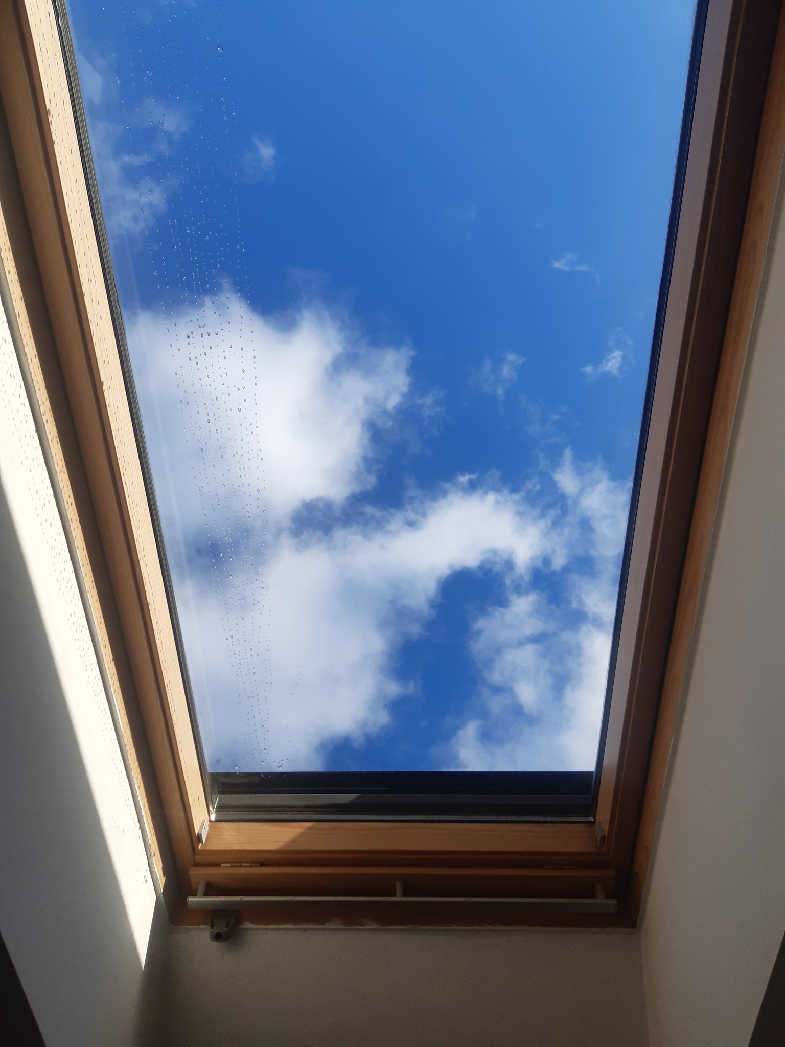 Okno dachowe z widokiem na błękitne niebo i białe chmury, krople wody na szybie po umyciu.