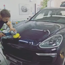 Mężczyzna poleruje maskę czarnego Porsche Cayenne w warsztacie, widoczne odbicie światła na lakierze, w tle regał z kosmetykami samochodowymi i obraz z sylwetką samochodu.