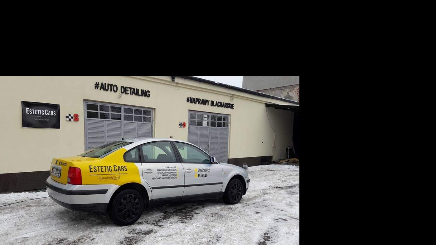 Srebrny samochód z żółtymi elementami karoserii zaparkowany przed budynkiem warsztatu z napisem #NAPRAWY BLACHARSKIE i #AUTO DETAILING nad bramami garażowymi, na ziemi leży śnieg.