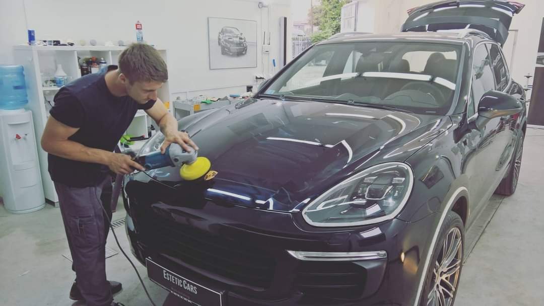 Mężczyzna poleruje maskę czarnego Porsche Cayenne w warsztacie, widoczne odbicie światła na lakierze, w tle regał z kosmetykami samochodowymi i obraz z sylwetką samochodu.