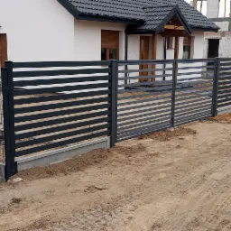 Nowoczesne ogrodzenie metalowe z poziomych paneli w kolorze grafitowym, oddzielające posesję z białym domem i budynkiem w trakcie budowy, widoczne ślady prac ziemnych.