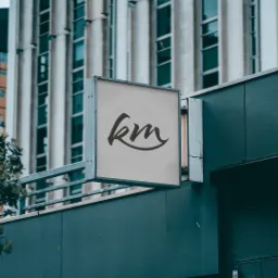 Szyld reklamowy z logo 'km' w minimalistycznym stylu, zamontowany na tle budynku z modernistyczną architekturą.