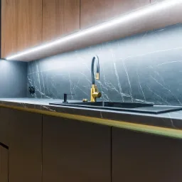 Nowoczesna kuchnia z czarnymi frontami, marmurowym blatem i złotą armaturą, oświetlona listwą LED pod szafkami górnymi.