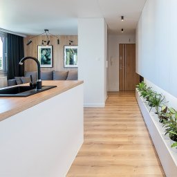 ASPEKT meble - Nowoczesne wnętrze mieszkania: wyspa kuchenna z drewnianym blatem i czarnym zlewem, widok na salon z szarą sofą i dekoracyjnymi plakatami, korytarz z zabudowaną szafą i roślinami w doniczkach.