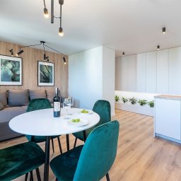 ASPEKT meble - Nowoczesne wnętrze mieszkania z drewnianą ścianą, szarą sofą, okrągłym białym stołem z zielonymi krzesłami, butelką wina i minimalistyczną białą zabudową kuchenną z roślinami.