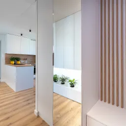 Nowoczesne wnętrze: biała kuchnia z drewnianymi akcentami, widok przez lustro na rośliny w minimalistycznej wnęce, drewniane lamele na ścianie.