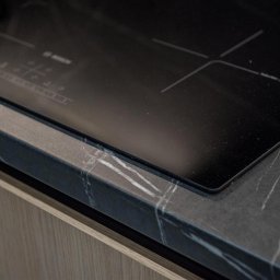 ASPEKT meble - Indukcyjna płyta grzewcza wbudowana w blat kuchenny z ciemnego marmuru z białymi żyłami, widoczny fragment frontu szafki z jasnego drewna.