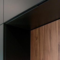 ASPEKT meble - Detal narożnika szafki z drewnopodobnym frontem, czarnym obramowaniem i szarą ścianą, widoczne logo producenta.