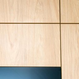 ASPEKT meble - Fragment frontu szafki z jasnego drewna z widocznym usłojeniem i granatowym, matowym wykończeniem dolnej krawędzi oraz logo producenta w prawym dolnym rogu.