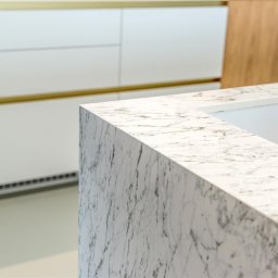 ASPEKT meble - Biały blat kuchenny z marmurowym wzorem, widoczne szafki z białymi frontami i złotymi akcentami, w tle ściana z drewnianą okleiną.