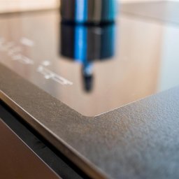 ASPEKT meble - Fragment blatu kuchennego z kamienną fakturą, widoczne krawędzie płyty indukcyjnej i logo producenta mebli w prawym dolnym rogu.