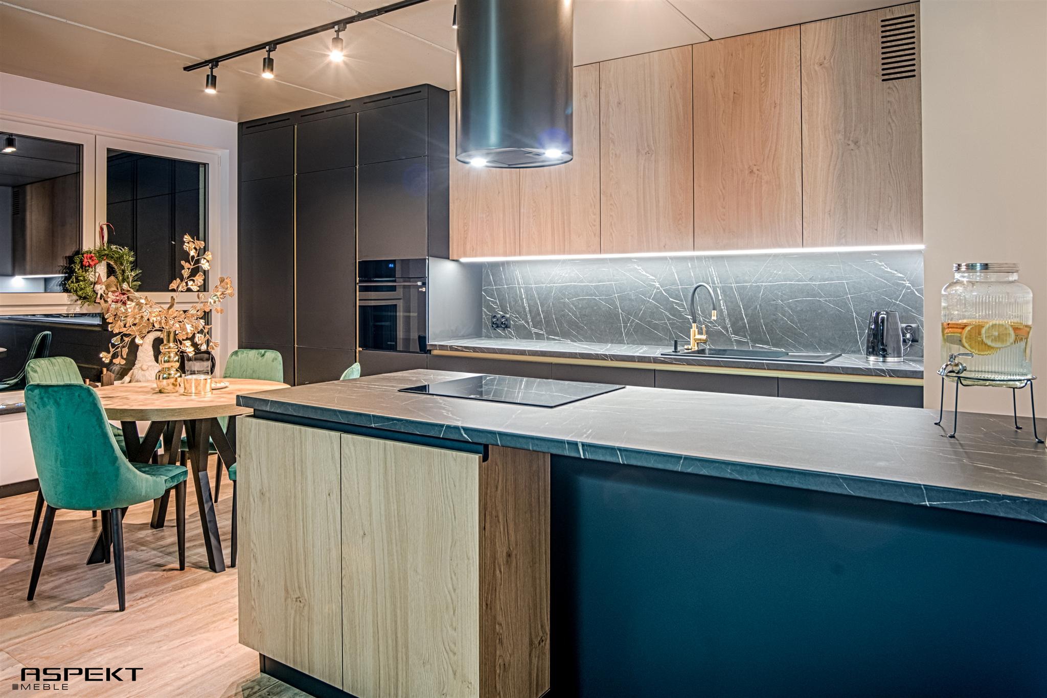 Nowoczesna kuchnia z wyspą, fronty szafek w odcieniach drewna i granatu, marmurowy backsplash, okrągły stół z zielonymi krzesłami, oświetlenie punktowe i dekoracje.