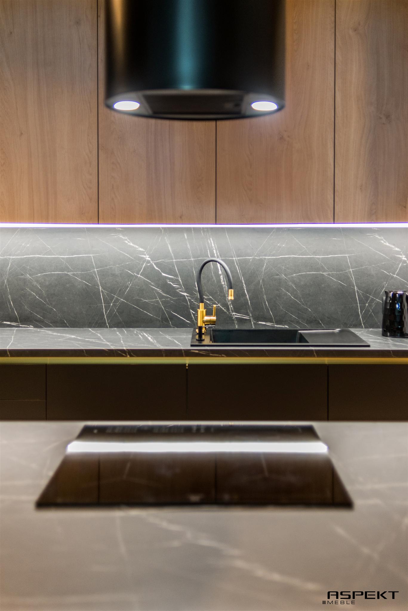 Nowoczesna kuchnia: drewniane szafki, marmurowy blat z czarnym zlewem i złotą baterią, oświetlenie LED pod szafkami, czarny okap.
