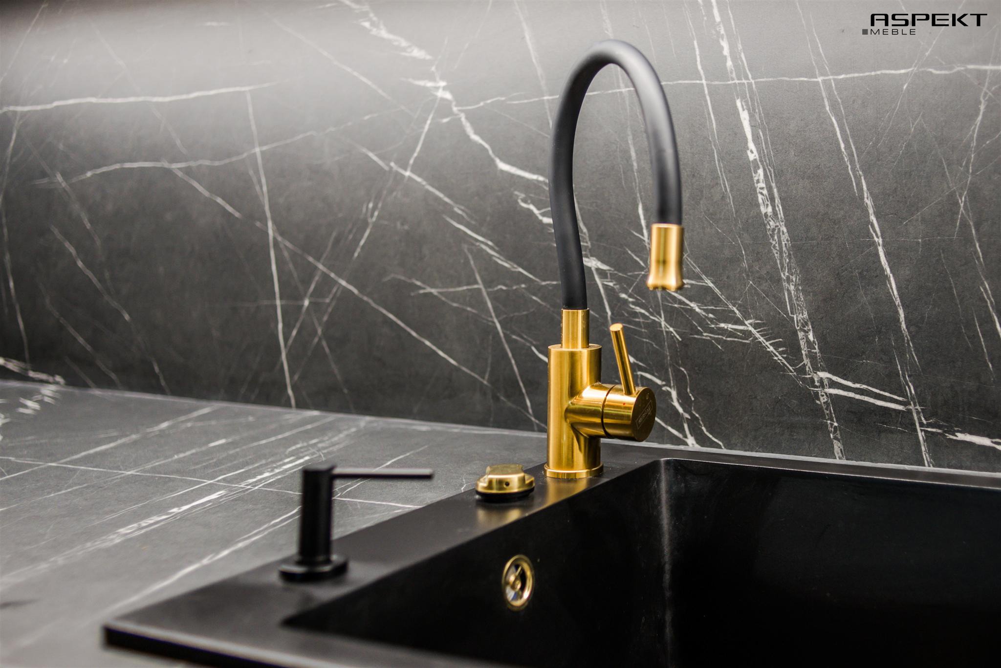 Czarny zlew z baterią w kolorze szczotkowanego złota i czarnym dozownikiem na tle blatu z czarnego marmuru z białymi żyłami oraz logo producenta mebli w prawym górnym rogu.