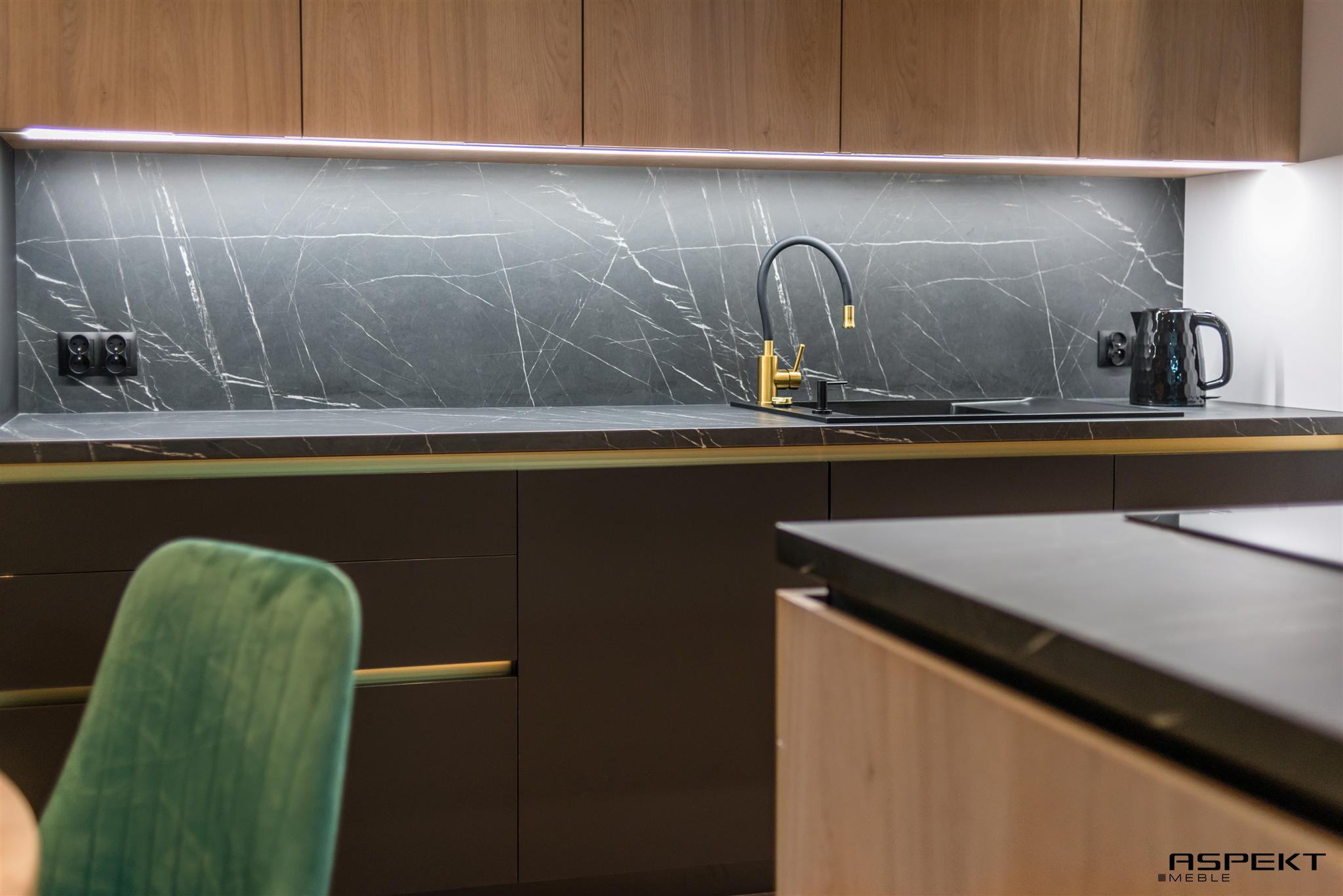 Nowoczesna kuchnia: ciemne, gładkie fronty szafek, blat z efektem marmuru, złota bateria i czajnik elektryczny, oświetlenie LED podszafkowe, zielone krzesło w tle.