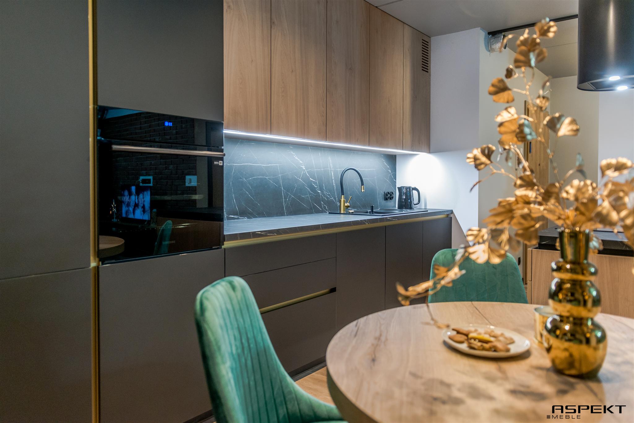 Nowoczesna kuchnia z szarymi matowymi frontami, drewnianymi górnymi szafkami, blatem z efektem marmuru oraz złotymi akcentami, w tym bateria i dekoracja z liści. Na stole okrągłe ciastka na talerzyku.