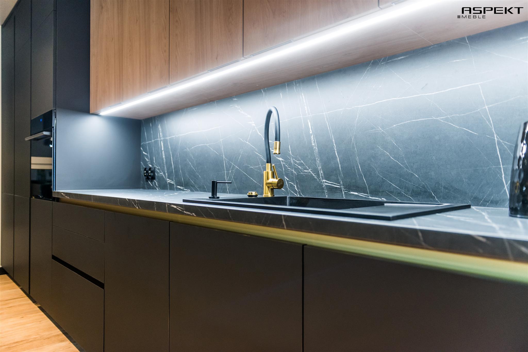 Nowoczesna kuchnia z czarnymi frontami, marmurowym blatem i złotą armaturą, oświetlona listwą LED pod szafkami górnymi.