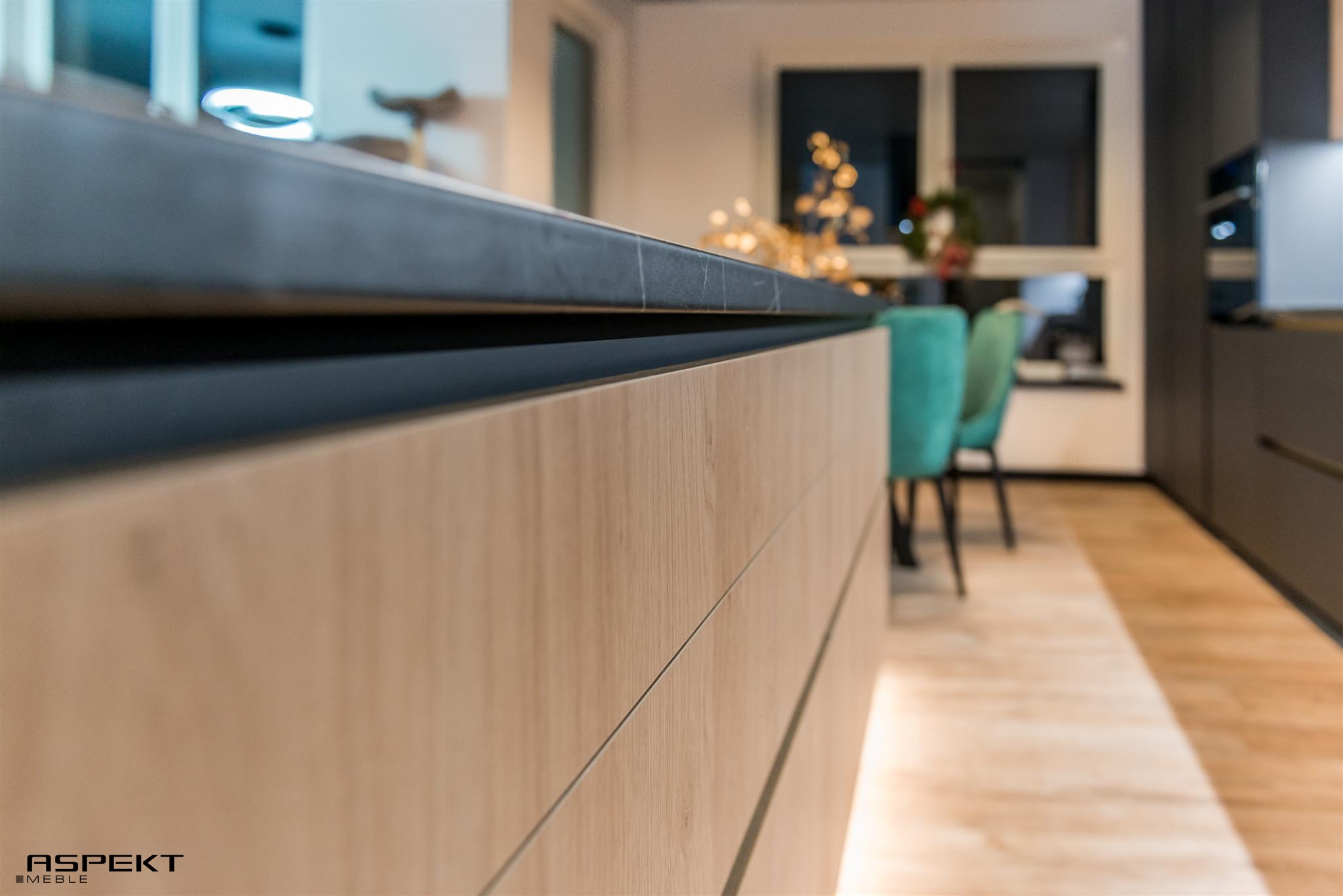 Detal blatu z ciemnego kompozytu i frontów szafek z jasnego drewna w nowoczesnej kuchni, z widocznym logo firmy 'Aspekt Meble' w lewym dolnym rogu.