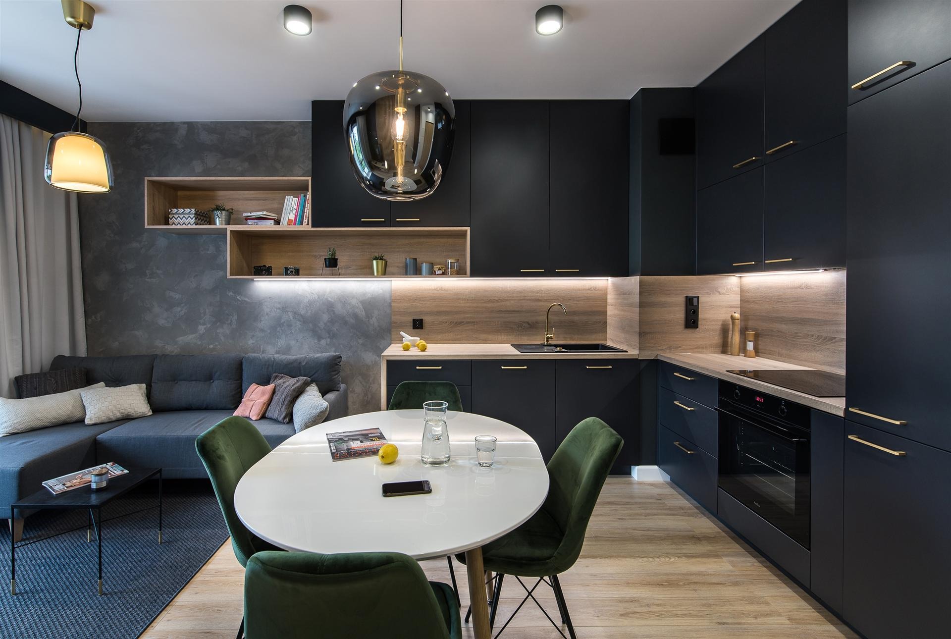 Nowoczesna kuchnia z czarnymi szafkami, drewnianym blatem, okrągłym białym stołem i zielonymi krzesłami w otwartej przestrzeni z szarą sofą i dekoracyjną ścianą.