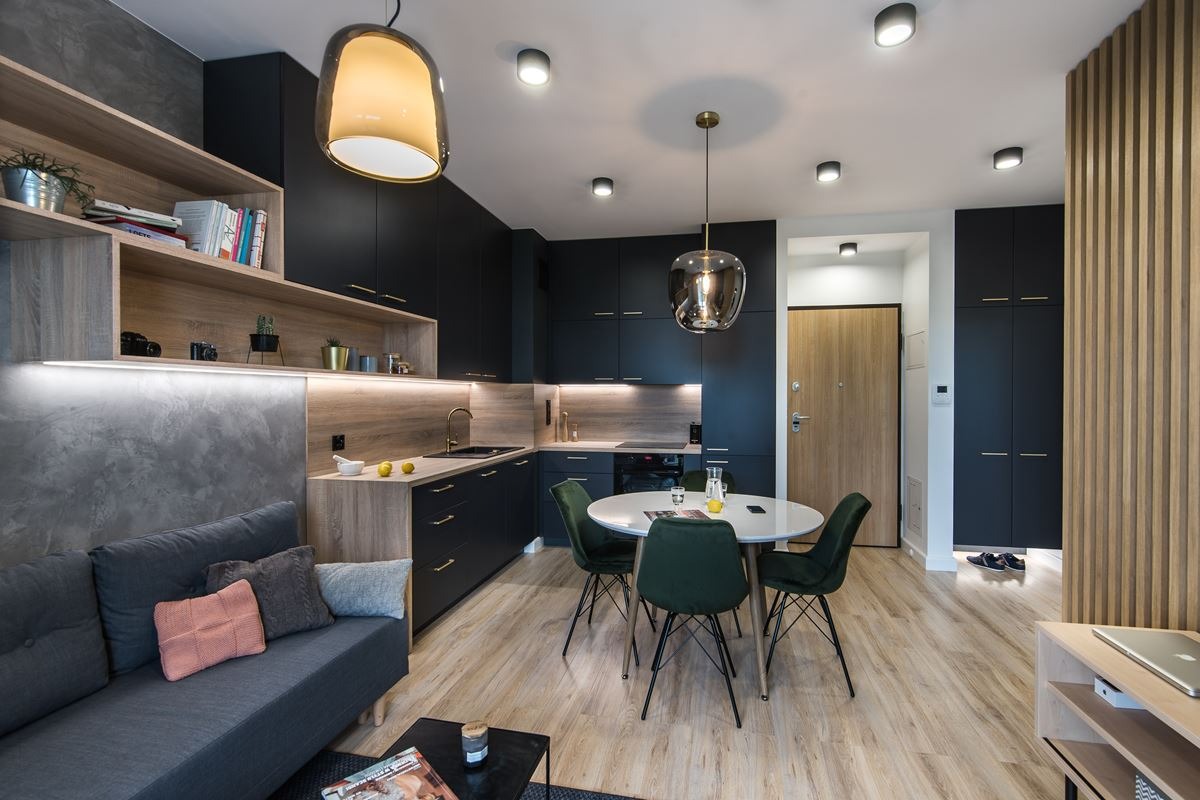 Nowoczesna aranżacja wnętrza: ciemne szafki kuchenne z drewnianymi akcentami, okrągły stół z zielonymi krzesłami, szara sofa z kolorowymi poduszkami i dekoracyjne oświetlenie.