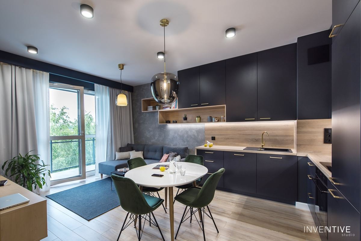 Nowoczesne wnętrze: ciemna zabudowa kuchenna z drewnianym panelem i złotą armaturą, okrągły biały stół z zielonymi krzesłami, szara sofa i balkon z widokiem na zieleń.