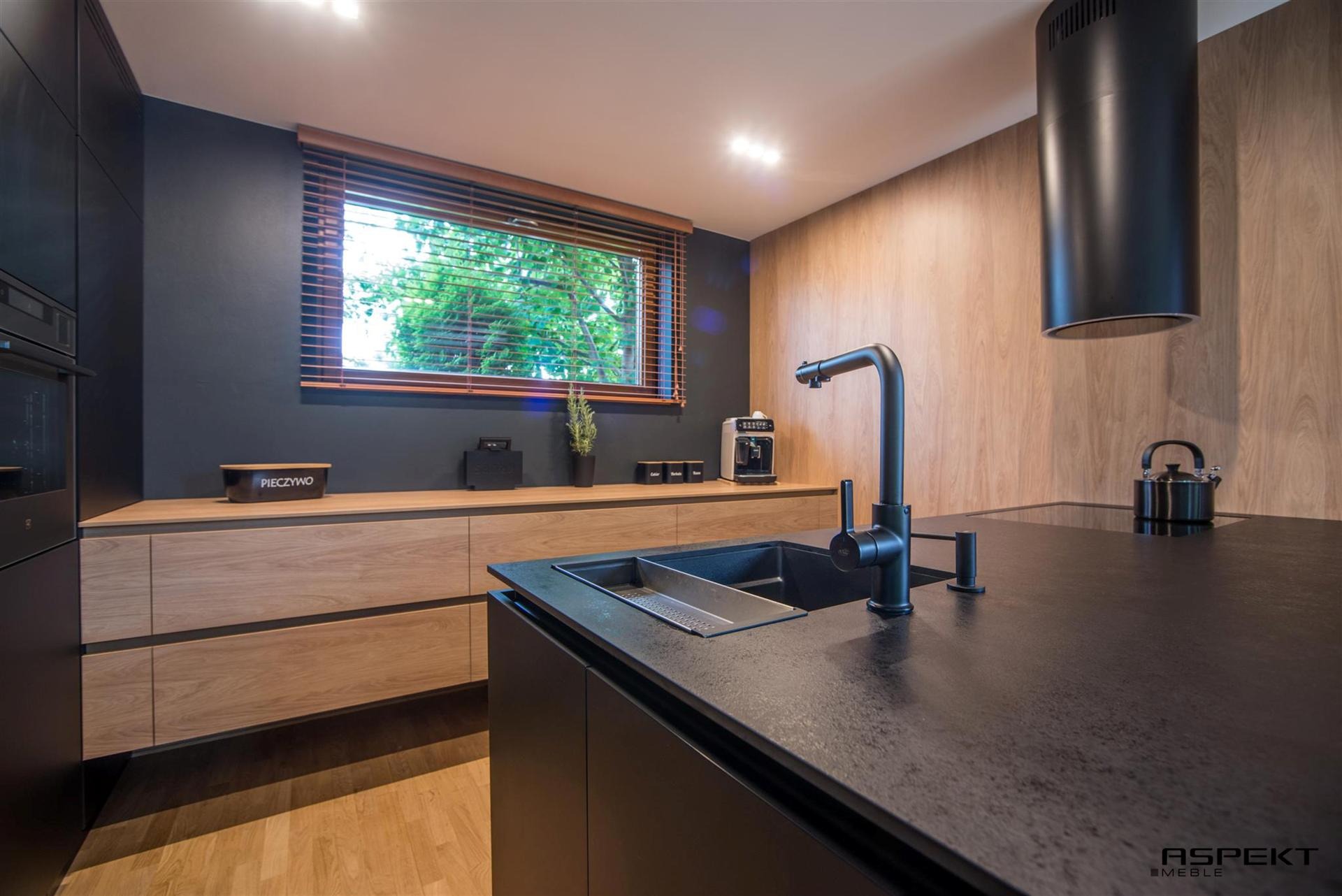 Nowoczesna kuchnia z czarnym blatem i armaturą, jasnymi drewnianymi frontami szafek, widok na okno z drewnianymi żaluzjami i pojemnikiem na pieczywo.