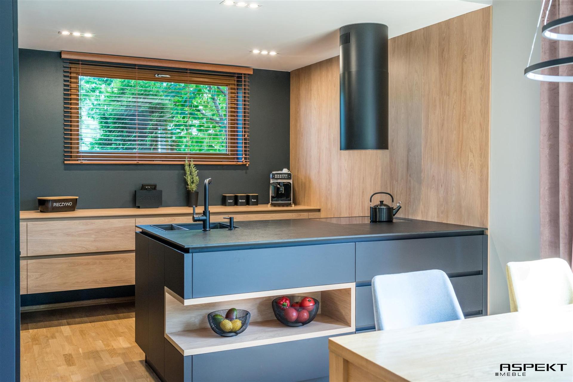 Nowoczesna kuchnia z szarymi szafkami, drewnianymi akcentami i czarnym okapem cylindrycznym, oświetlona światłem dziennym wpadającym przez okno z żaluzjami.