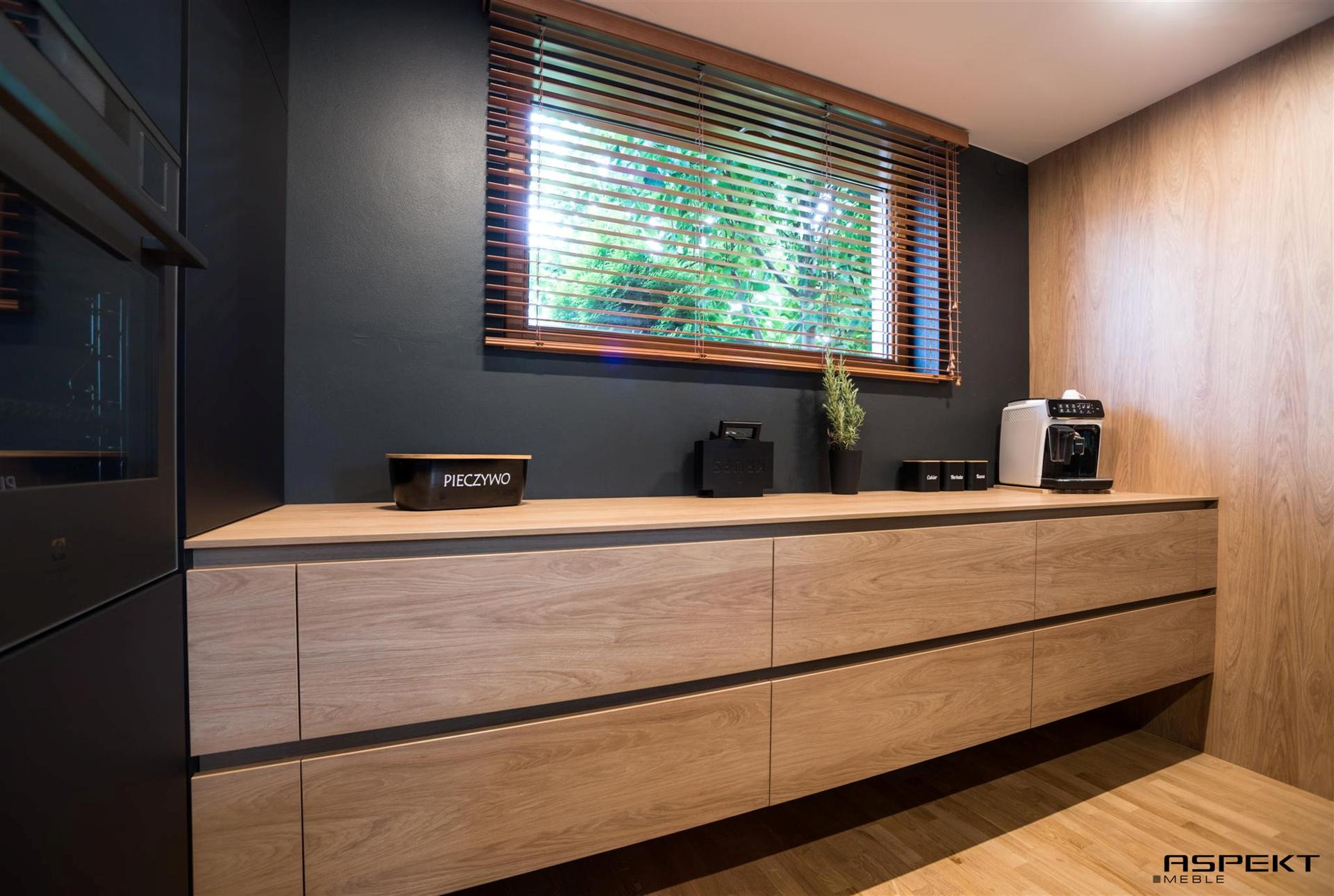 Nowoczesna kuchnia: drewniane fronty szafek z subtelnymi, ciemnymi wstawkami, blaty z akcesoriami kuchennymi i oknem z drewnianymi żaluzjami, na tle ciemnej ściany.