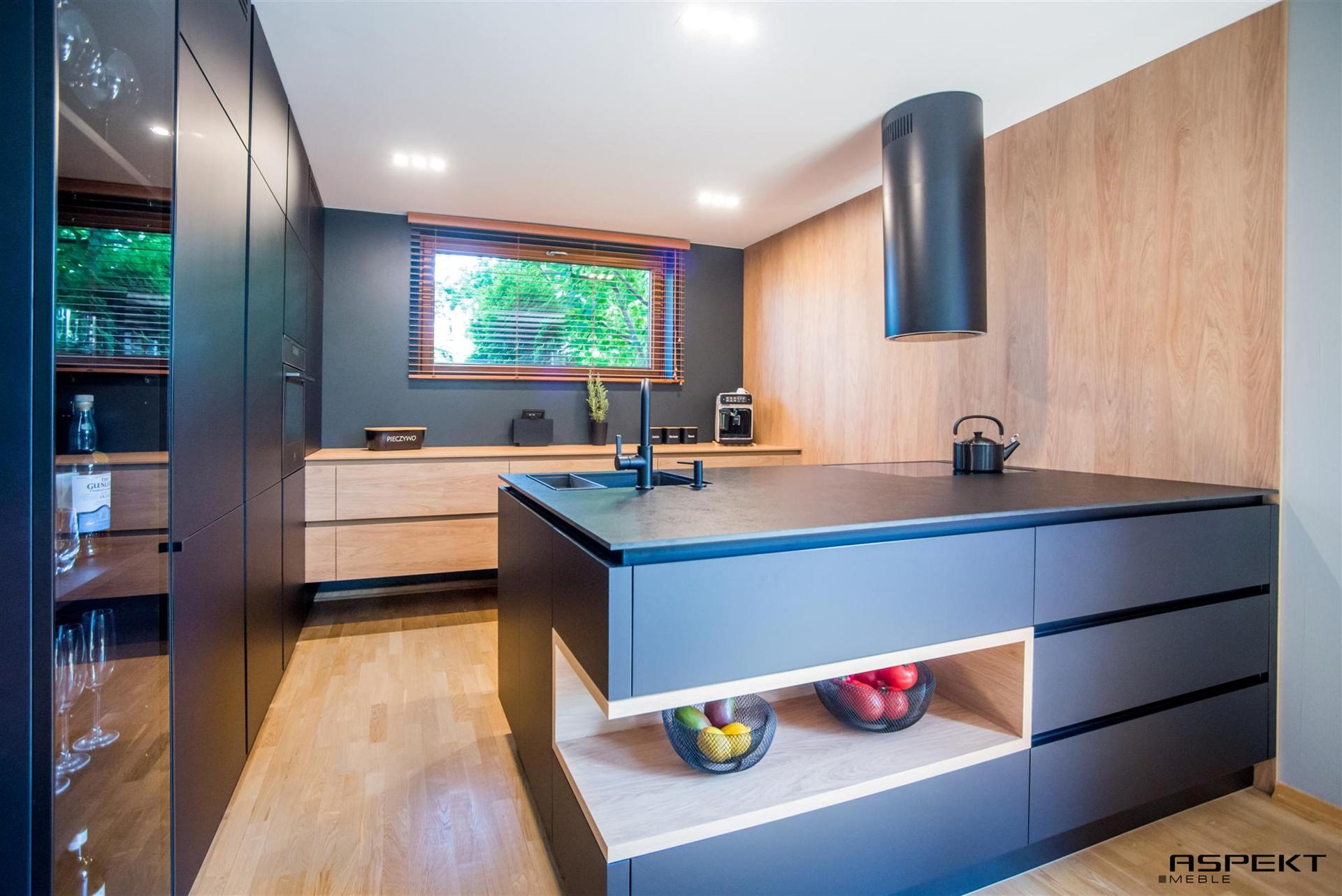 Nowoczesna kuchnia z wyspą w odcieniach szarości i drewna, czarne matowe fronty szafek, drewniane blaty, oświetlenie punktowe, wyspa z otwartymi półkami na owoce w metalowych koszykach, okno...