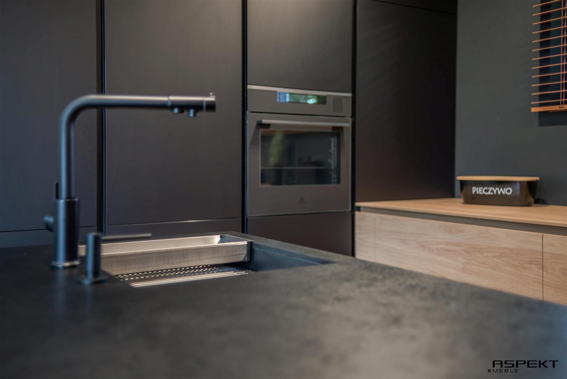 Minimalistyczna kuchnia w ciemnych odcieniach szarości i czerni, z matowymi frontami szafek, nowoczesną armaturą i wbudowanym piekarnikiem. Na blacie widoczny pojemnik na pieczywo.