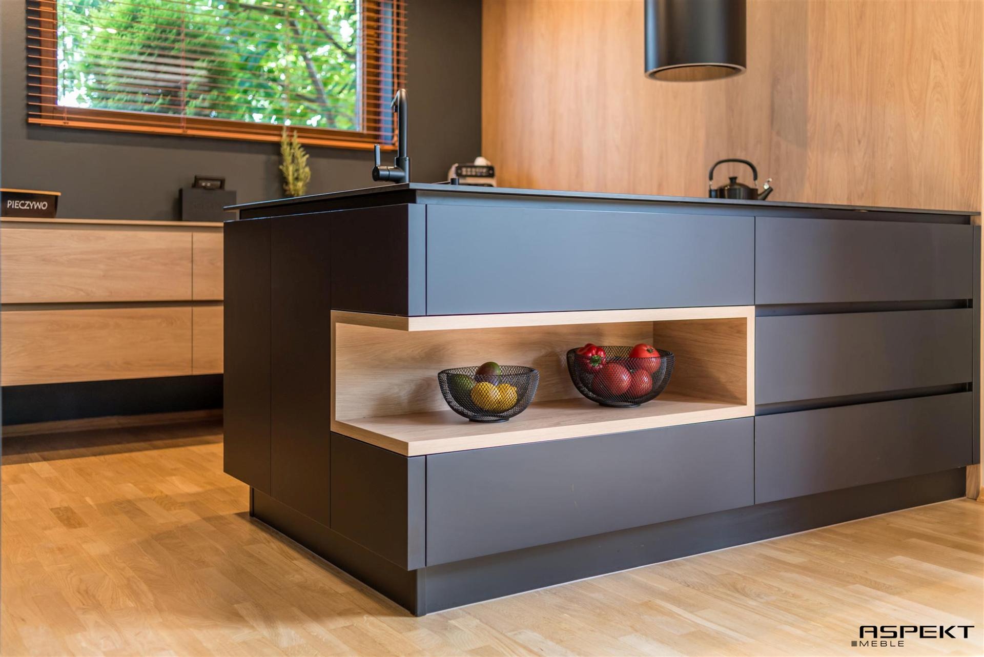 Nowoczesna wyspa kuchenna z szarymi frontami, drewnianym blatem i otwartą półką z koszykami owoców, na tle drewnianej ściany i okna z żaluzjami.