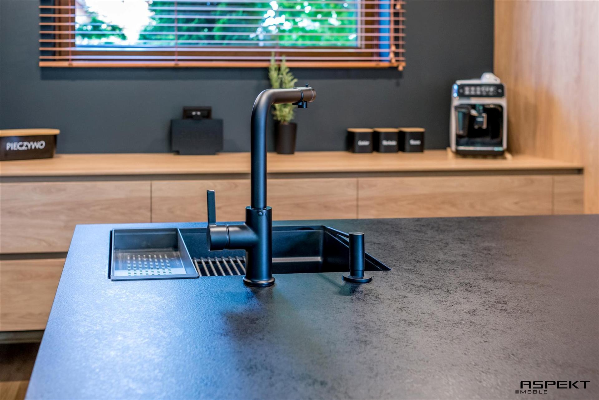 Czarny, matowy kran i zlew wkomponowany w ciemny blat z szorstką fakturą, w tle drewniane szafki i okno z żaluzjami.