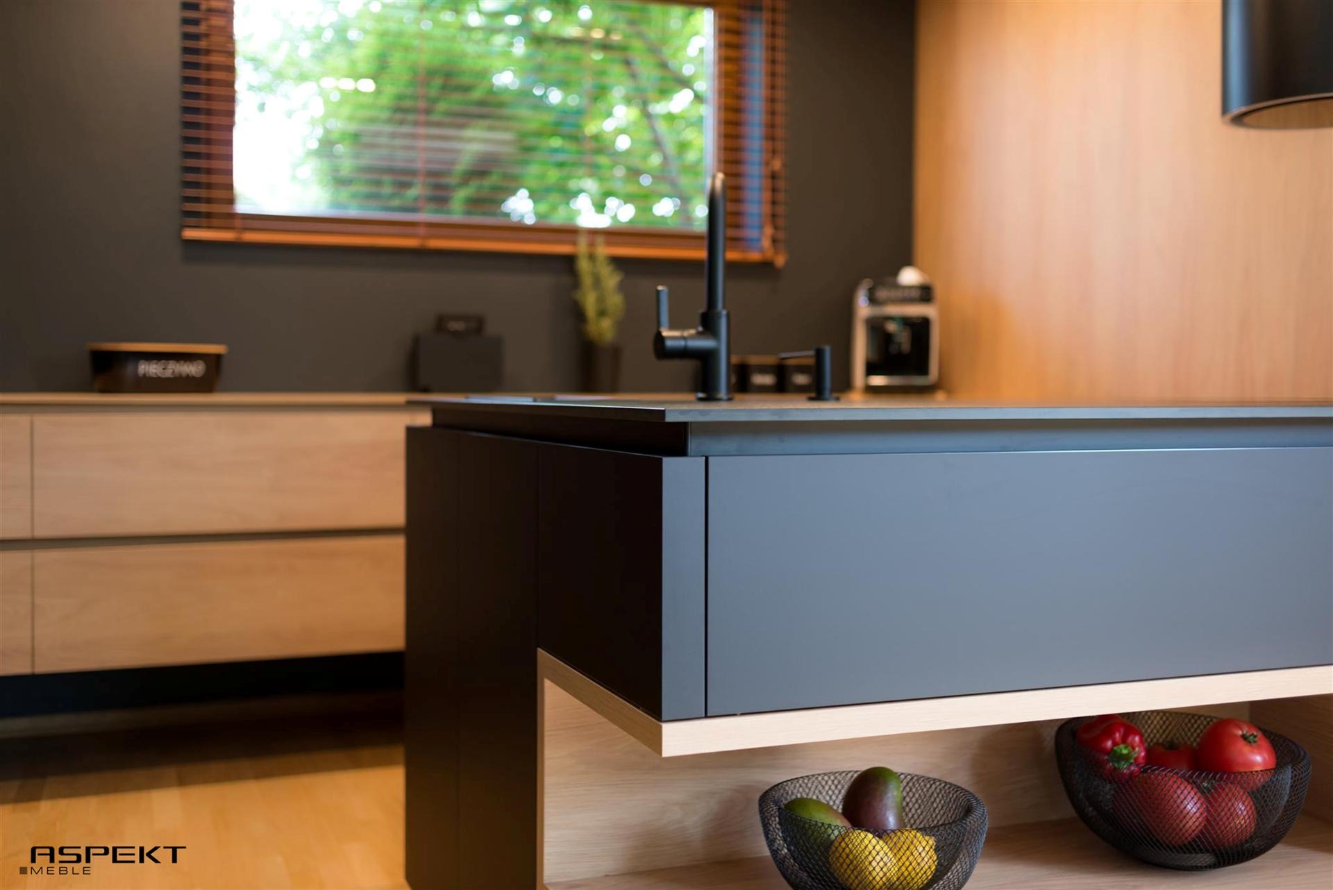 Nowoczesna wyspa kuchenna z szarymi frontami i drewnianymi akcentami, czarny kran, koszyki z owocami i warzywami, jasne drewno w tle, widok z boku.
