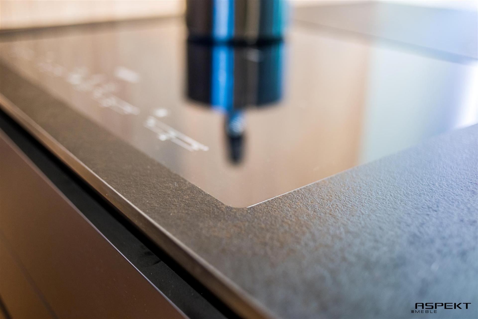 Fragment blatu kuchennego z kamienną fakturą, widoczne krawędzie płyty indukcyjnej i logo producenta mebli w prawym dolnym rogu.