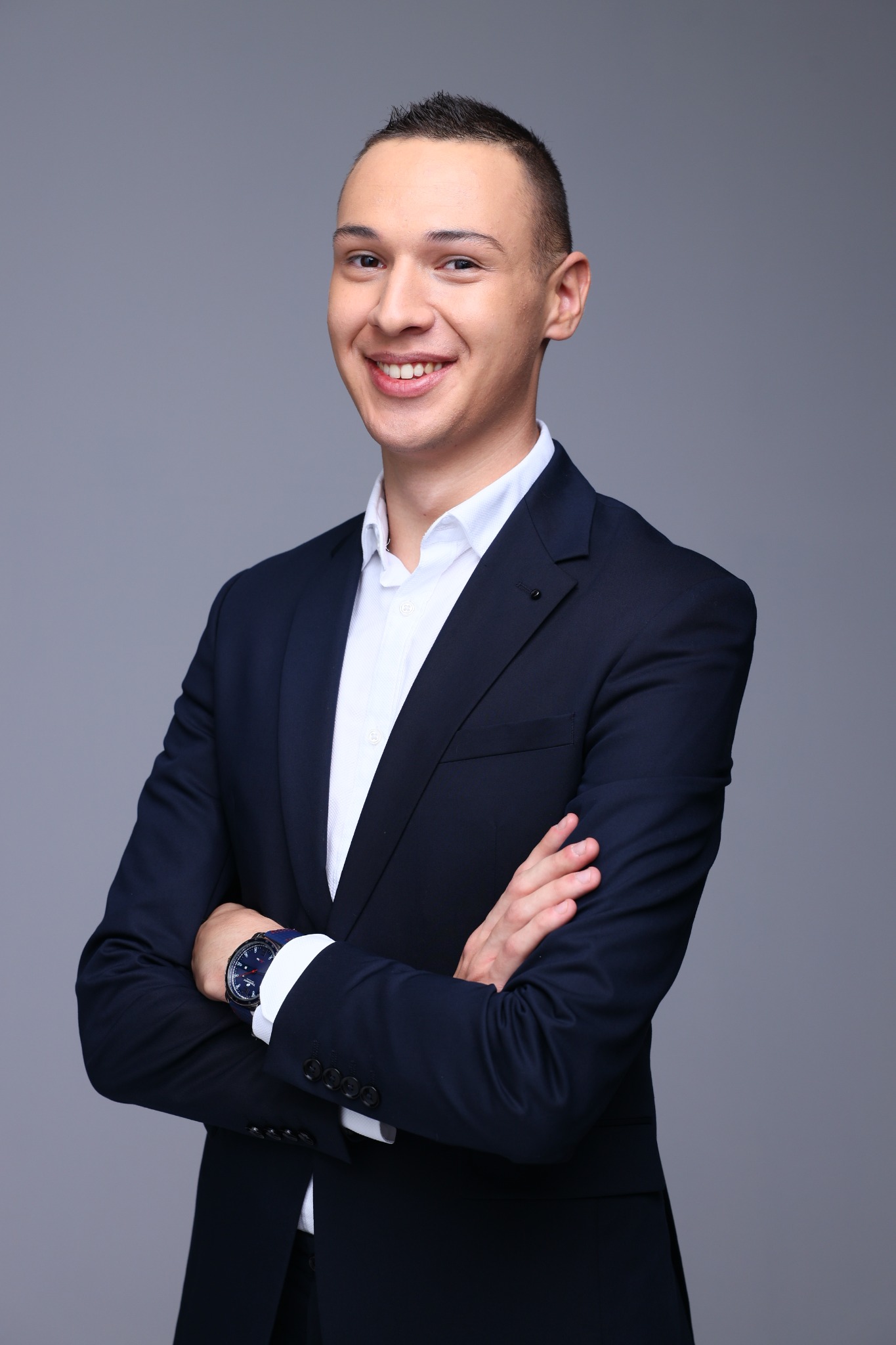 Uśmiechnięty młody mężczyzna w granatowym garniturze i białej koszuli, z zegarkiem na nadgarstku, pozuje na szarym tle z założonymi rękami.