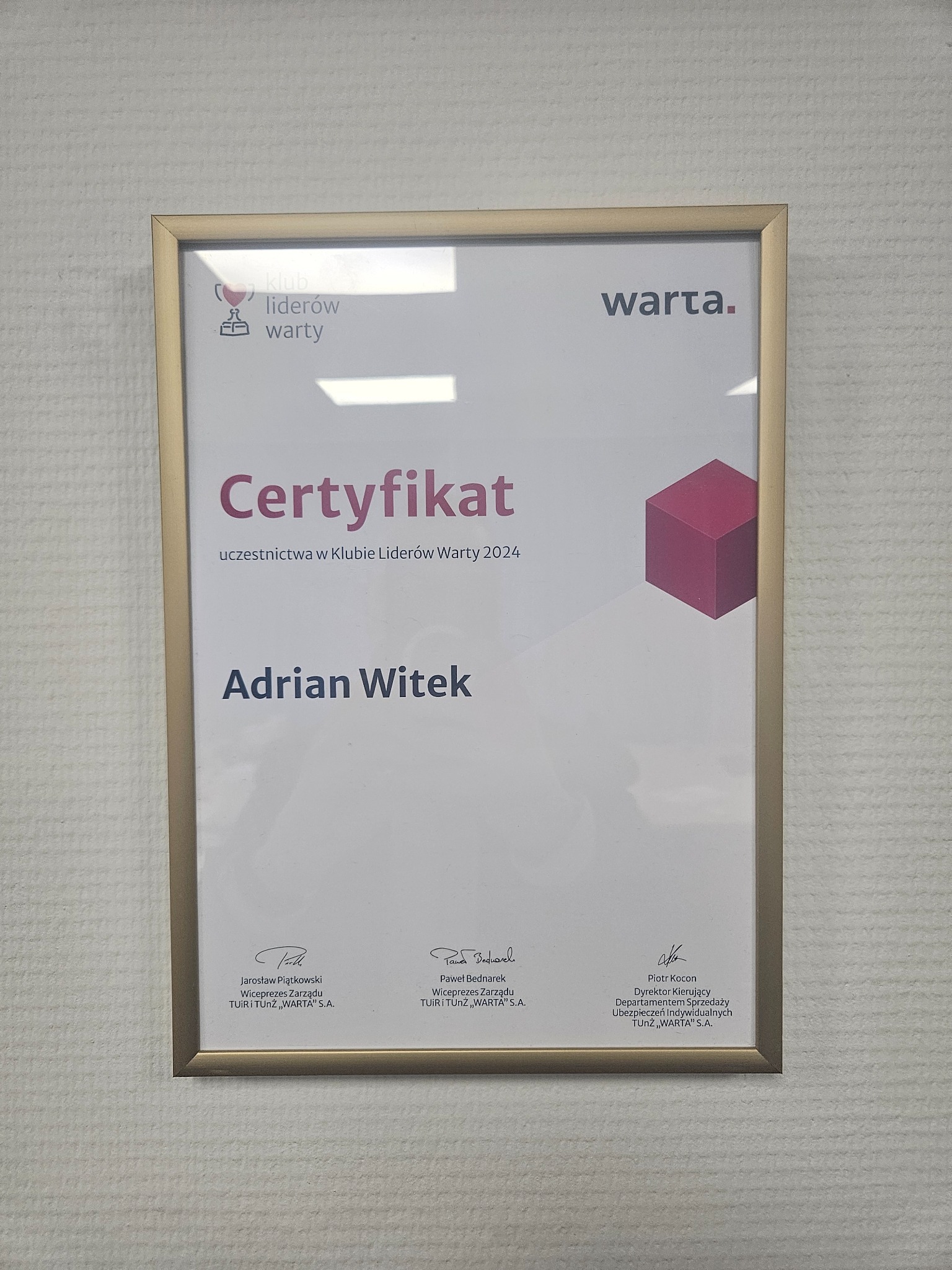 Oprawiony w złotą ramkę certyfikat Klubu Liderów Warty dla Adriana Witka, wiszący na jasnej ścianie.