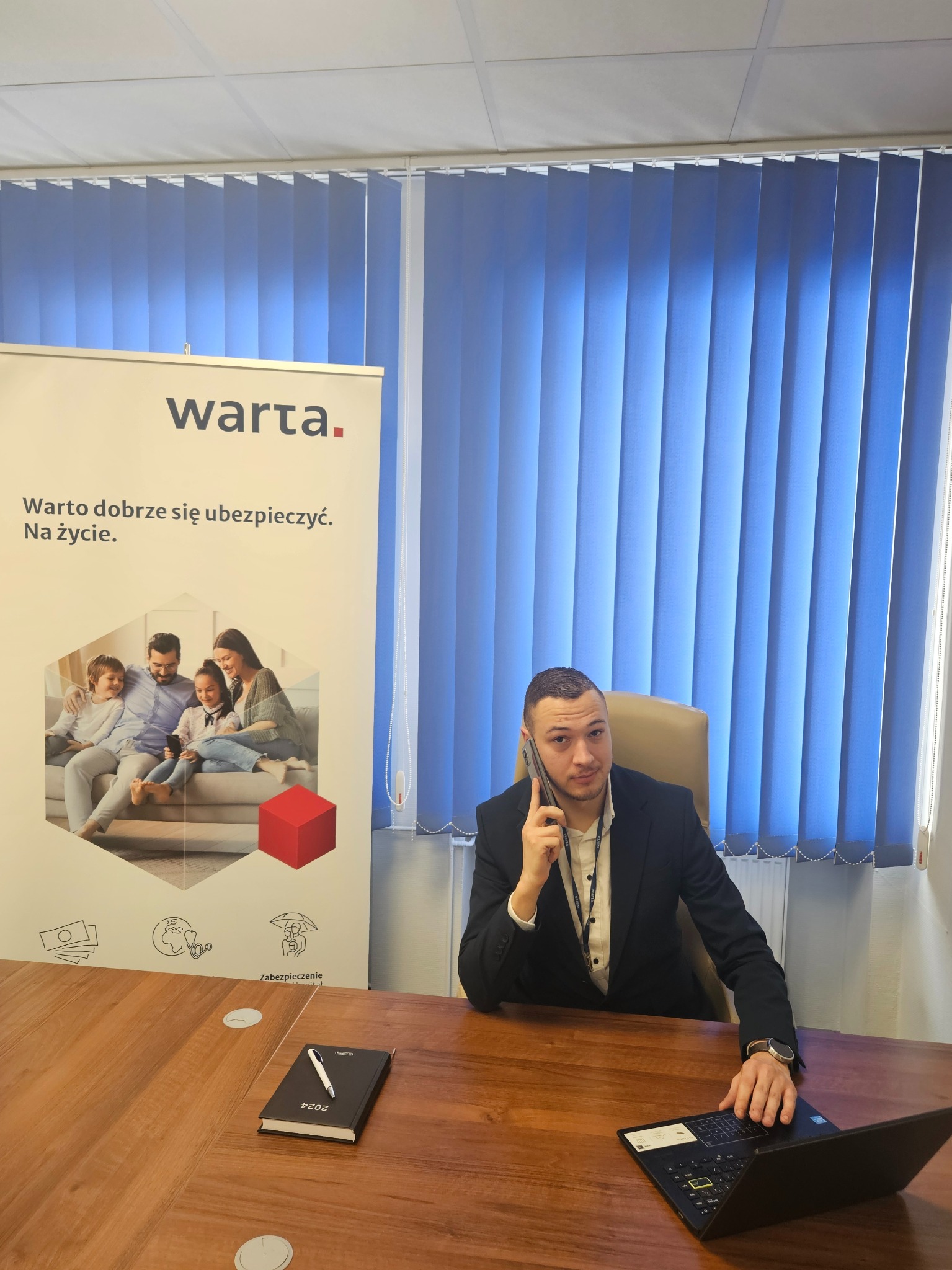 Agent ubezpieczeniowy Warta rozmawia przez telefon w biurze, obok baner reklamowy z rodziną i hasłem 'Warto dobrze się ubezpieczyć. Na życie.'
