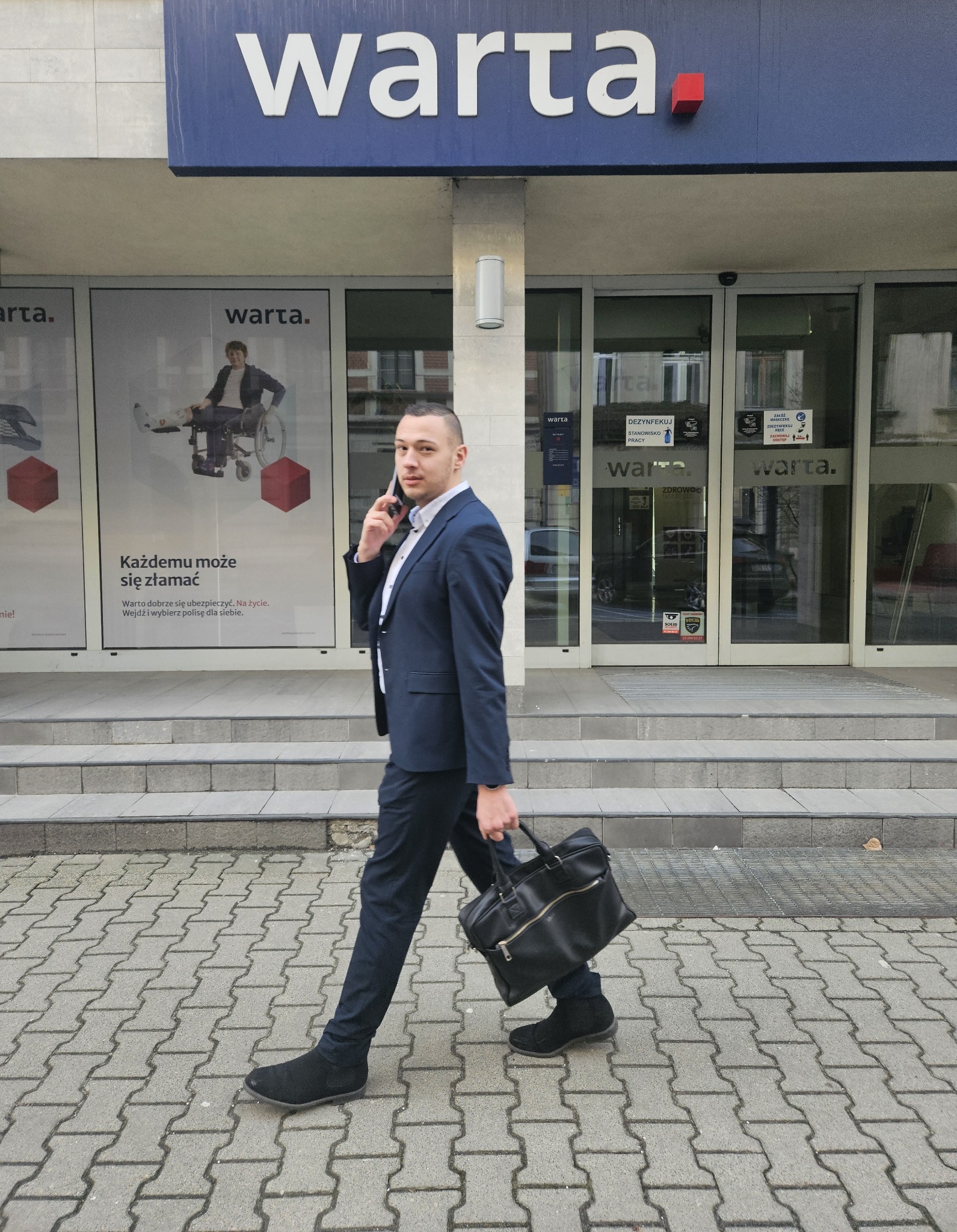 Mężczyzna w garniturze rozmawia przez telefon, trzymając czarną torbę, na tle biura Warta z reklamami ubezpieczeń.