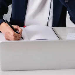 Osoba w garniturze pisze czarnym długopisem w notatniku na biurku z laptopem, dokumentami i flagami Brazylii, Wielkiej Brytanii i USA w tle.