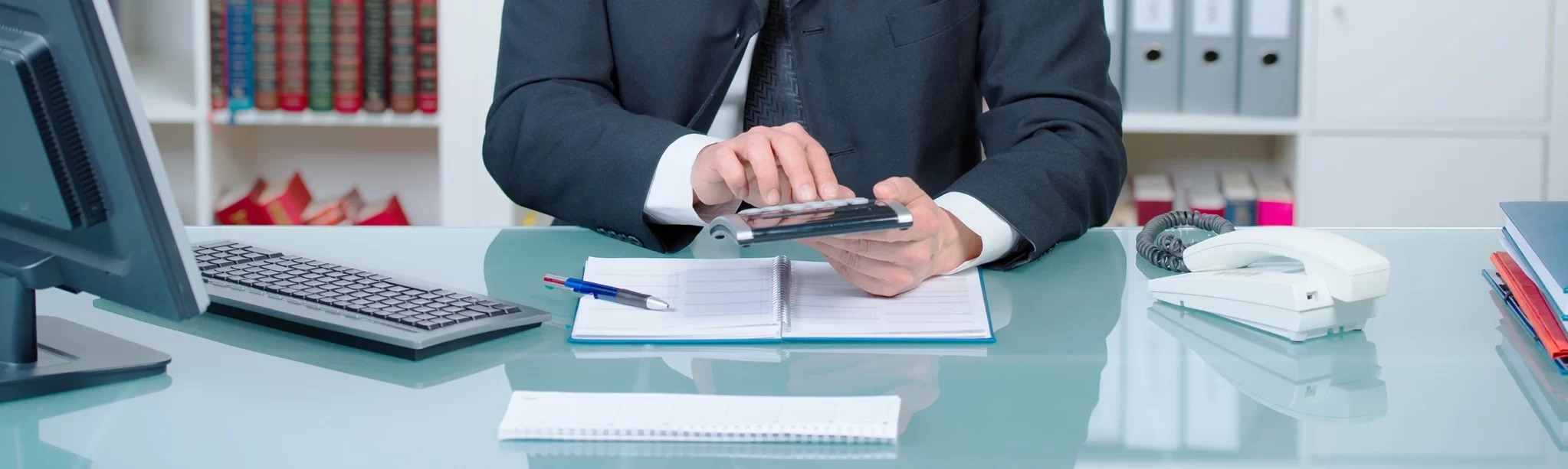 Osoba w garniturze wpisuje dane na kalkulatorze nad otwartym notatnikiem, na biurku telefon stacjonarny i monitor komputera.