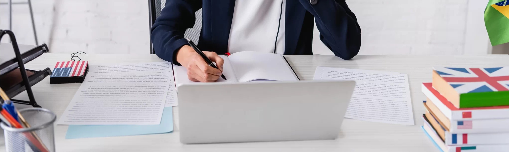 Osoba w garniturze pisze czarnym długopisem w notatniku na biurku z laptopem, dokumentami i flagami Brazylii, Wielkiej Brytanii i USA w tle.