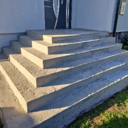 Nowe, betonowe schody wejściowe prowadzące do domu z nowoczesnymi drzwiami. Widoczne świeże wykończenie i detale architektoniczne. Słoneczny dzień.