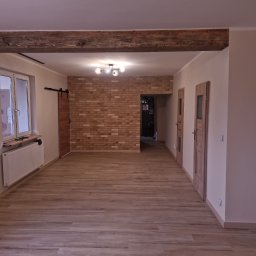 F.Bud - Wykończone wnętrze: drewniana podłoga, ściana z cegły, przesuwne drzwi stodołowe, drewniana belka stropowa i nowoczesna lampa sufitowa. Widok na sąsiednie pomieszczenie.