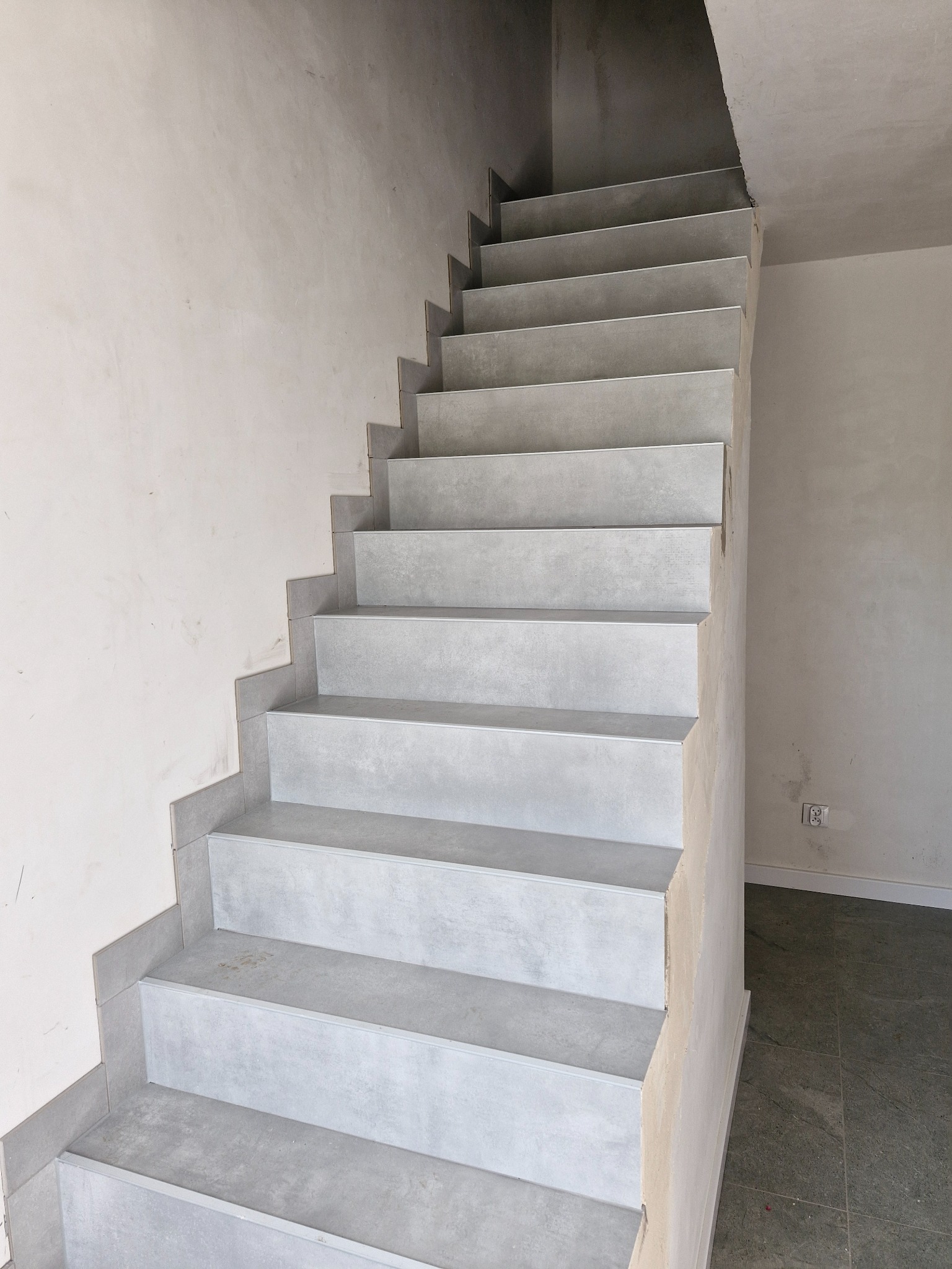 Wykończone schody wewnętrzne wyłożone szarymi płytkami, fragment ściany przygotowanej do malowania, widoczny kontakt elektryczny.