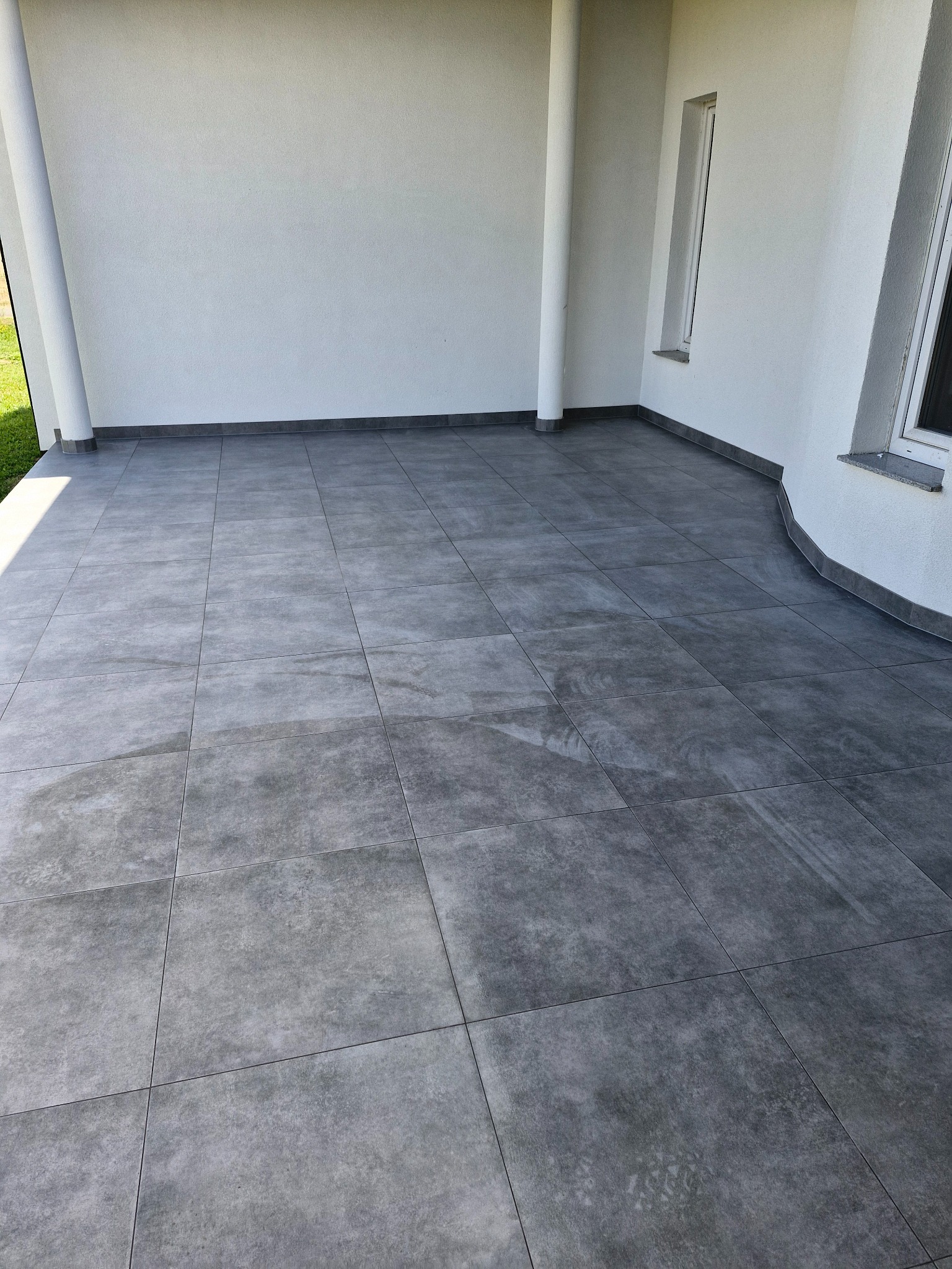 Wyłożony szarymi płytkami taras z widocznymi kolumnami podtrzymującymi zadaszenie i oknem w ścianie. Płytki ułożone równo, z widocznymi fugami. Minimalistyczny styl.