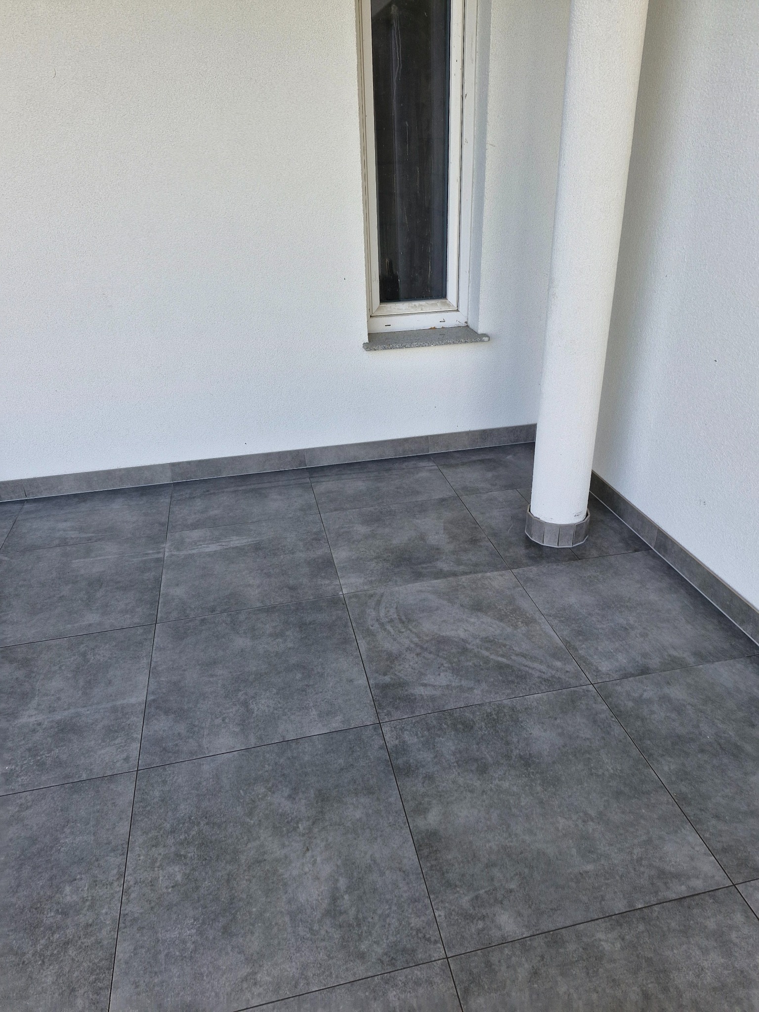 Wykończony taras w Trzcinnej z szarymi płytkami gresowymi o matowym wykończeniu, kolumna nośna w rogu oraz okno z białymi ramami.