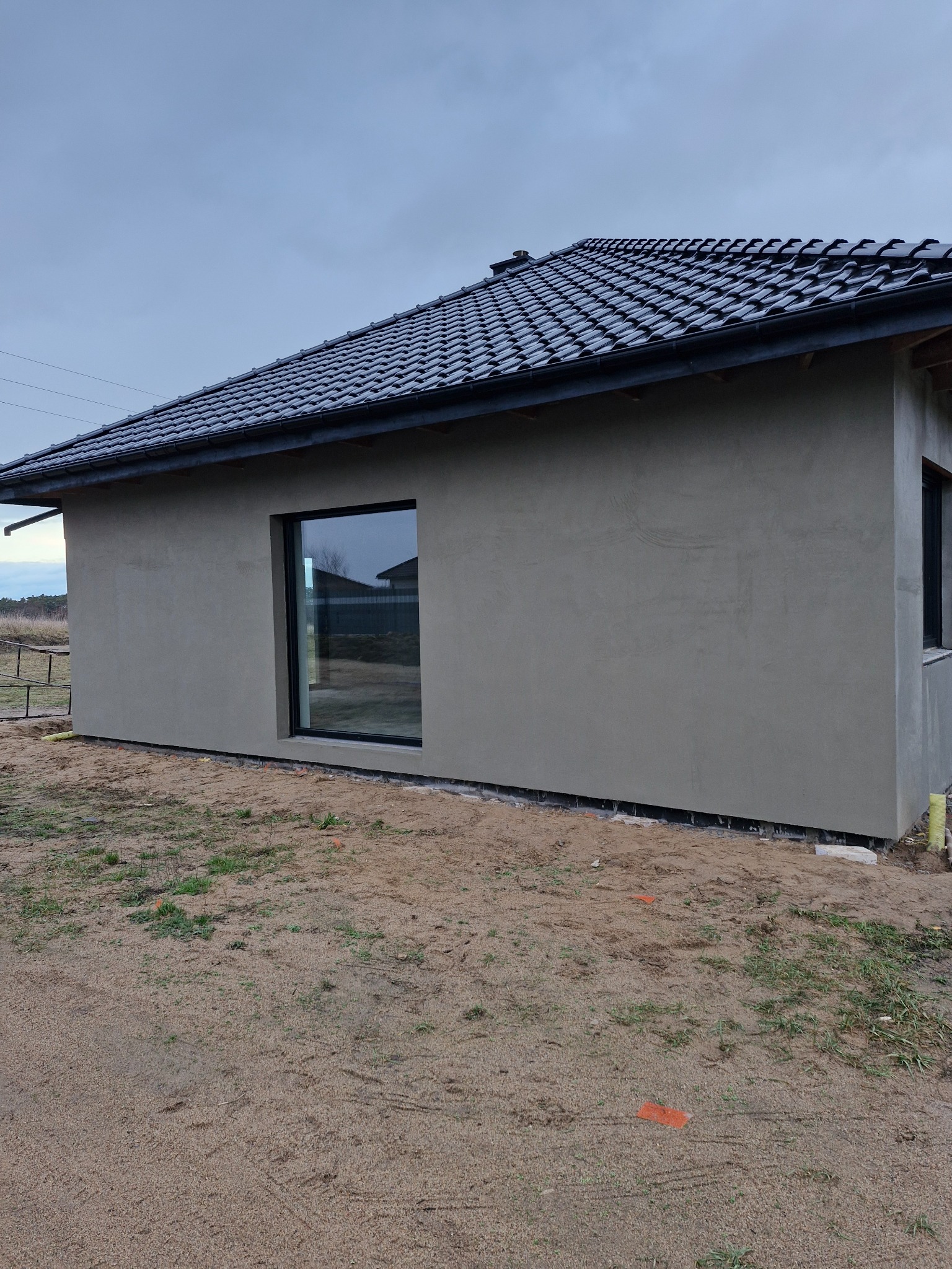 Surowy budynek z oknem w czarnej ramie, otynkowany szarą zaprawą, z ciemną dachówką, na piaszczystym terenie z pomarańczowymi znacznikami geodezyjnymi.